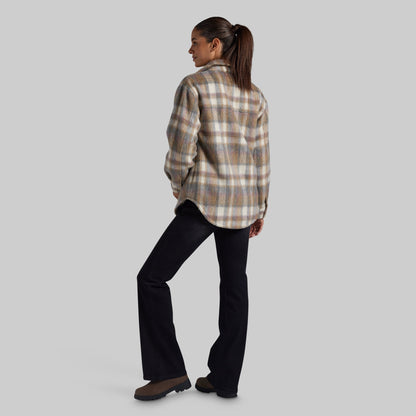 A woman is shown from the back wearing a beige and brown plaid flannel jacket and black pants, standing against a solid gray studio background.
