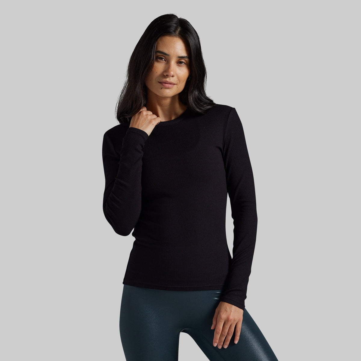 A woman modeling a black long-sleeve crewneck top against a solid gray background.