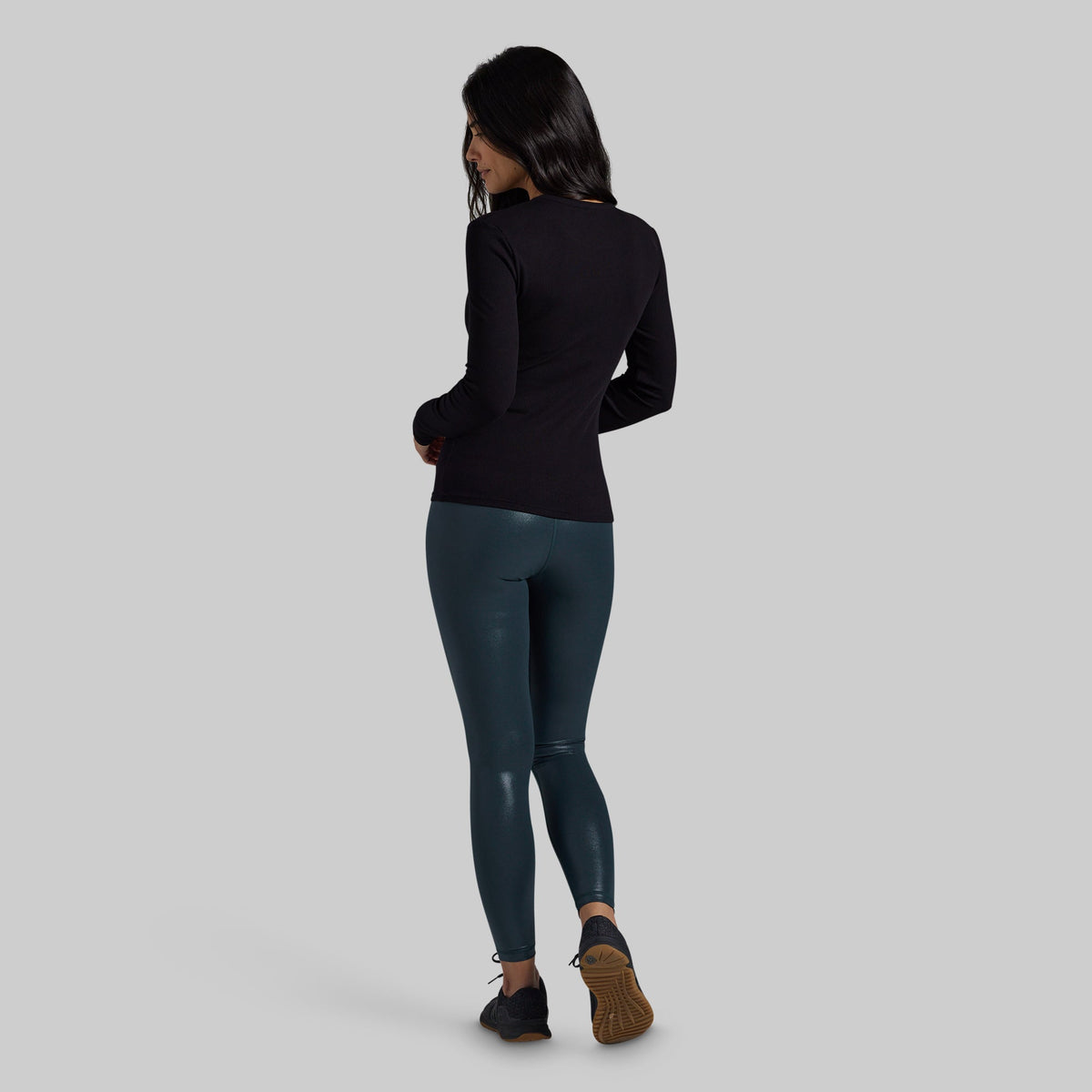 A woman is shown from the back wearing a black long-sleeve top and dark teal shiny leggings, standing against a solid light gray background.