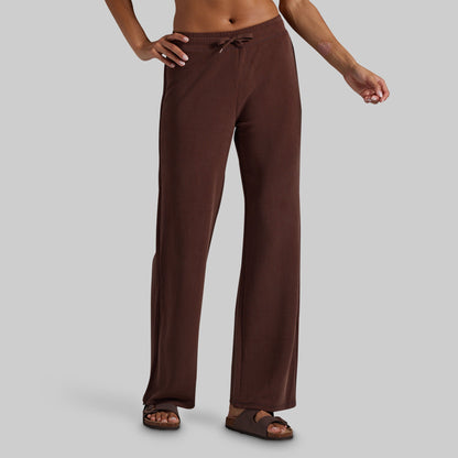 A person wearing loose brown drawstring lounge pants and brown sandals, shown from the waist down against a light gray studio background.