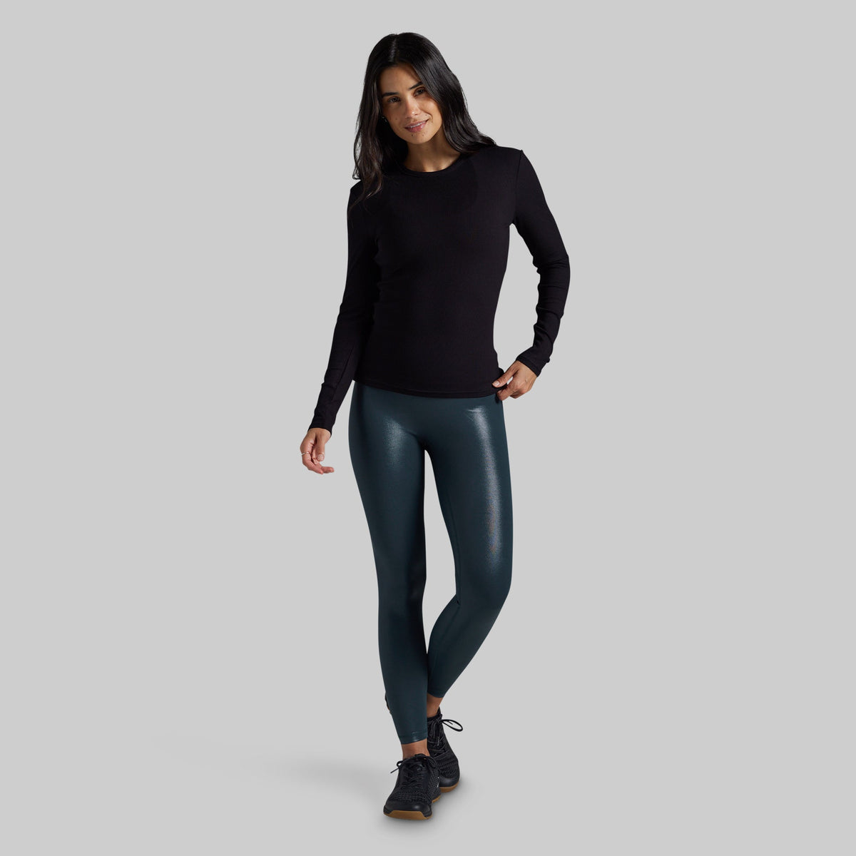 A woman modeling a black long-sleeve crewneck top and dark teal faux-leather leggings with black sneakers, standing against a solid gray studio background.