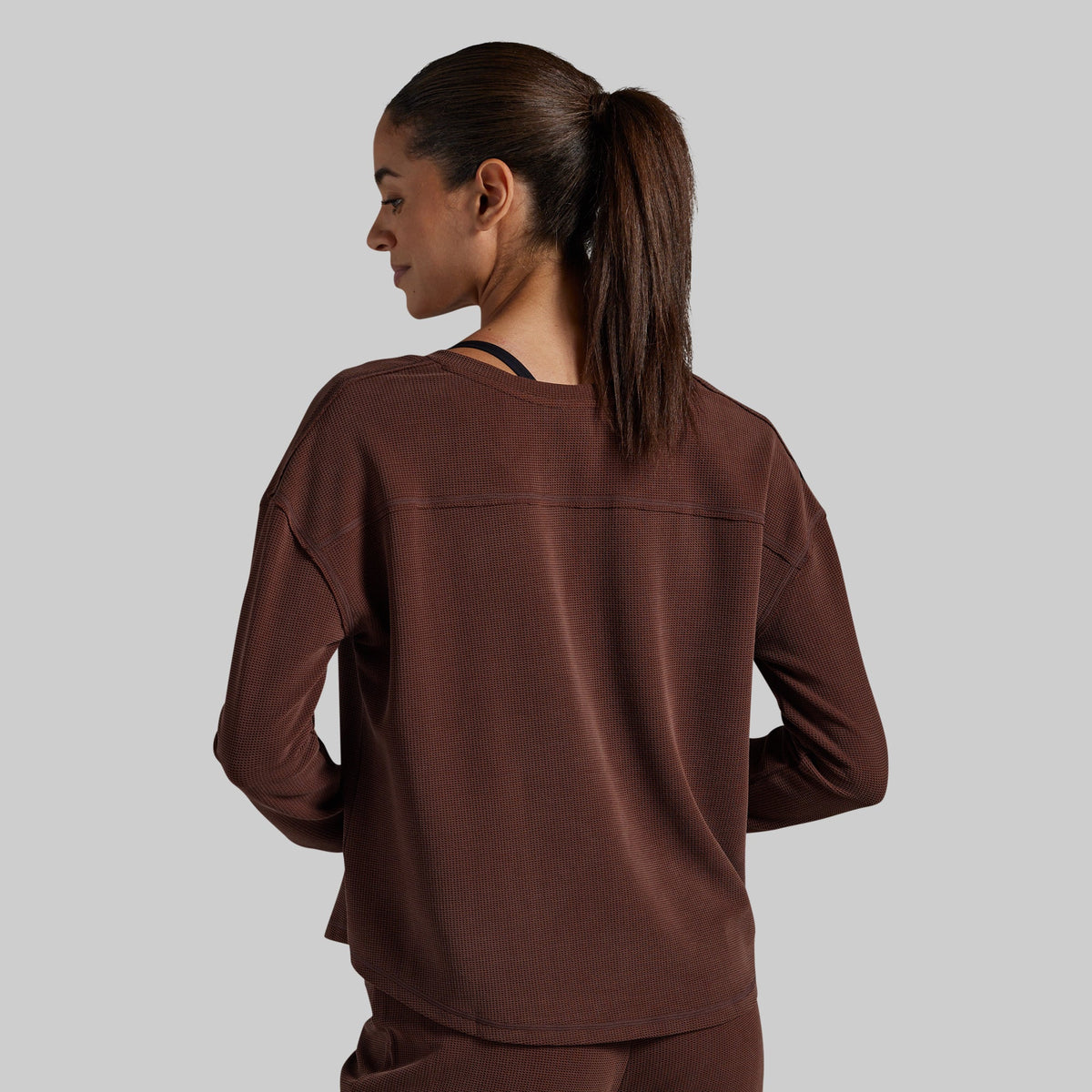 A woman is shown from the back wearing a dark brown long-sleeve top and matching pants against a solid gray background.
