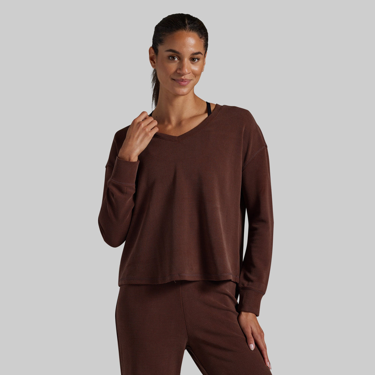 A woman modeling a matching brown lounge set consisting of a long-sleeve cropped V-neck top and coordinating pants, photographed against a solid light gray background.
