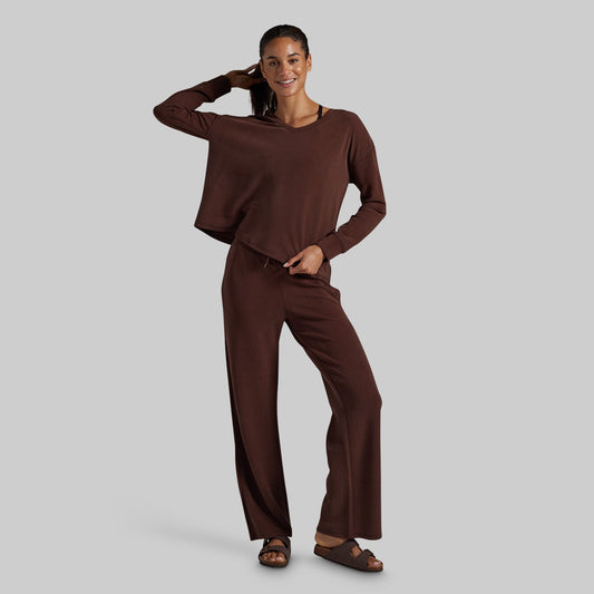 A woman modeling a matching dark brown lounge set consisting of a long-sleeve cropped top and wide-leg pants, paired with brown sandals, against a solid light gray background.