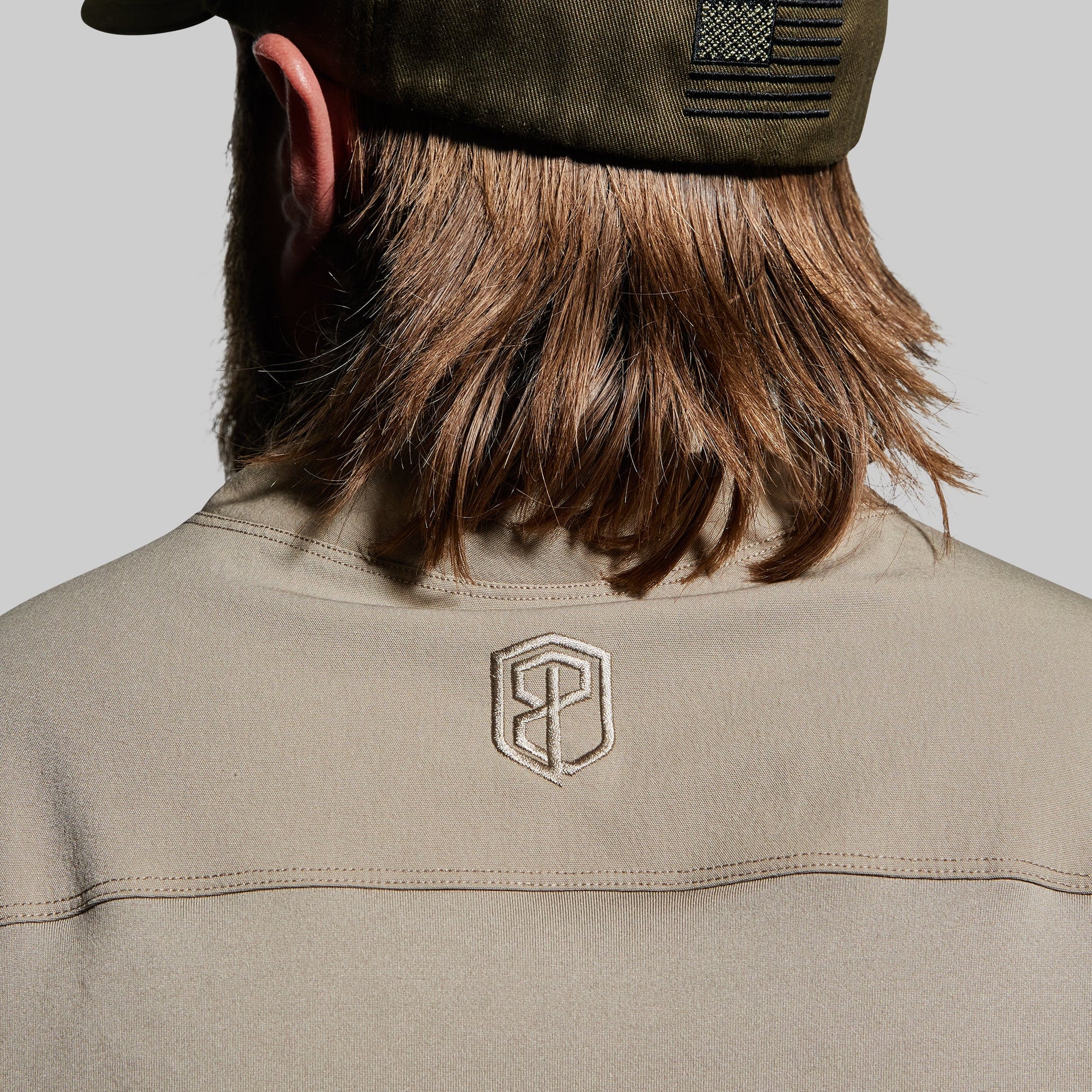 Person wearing a beige tactical shirt with a logo on the back, looking over their shoulder.