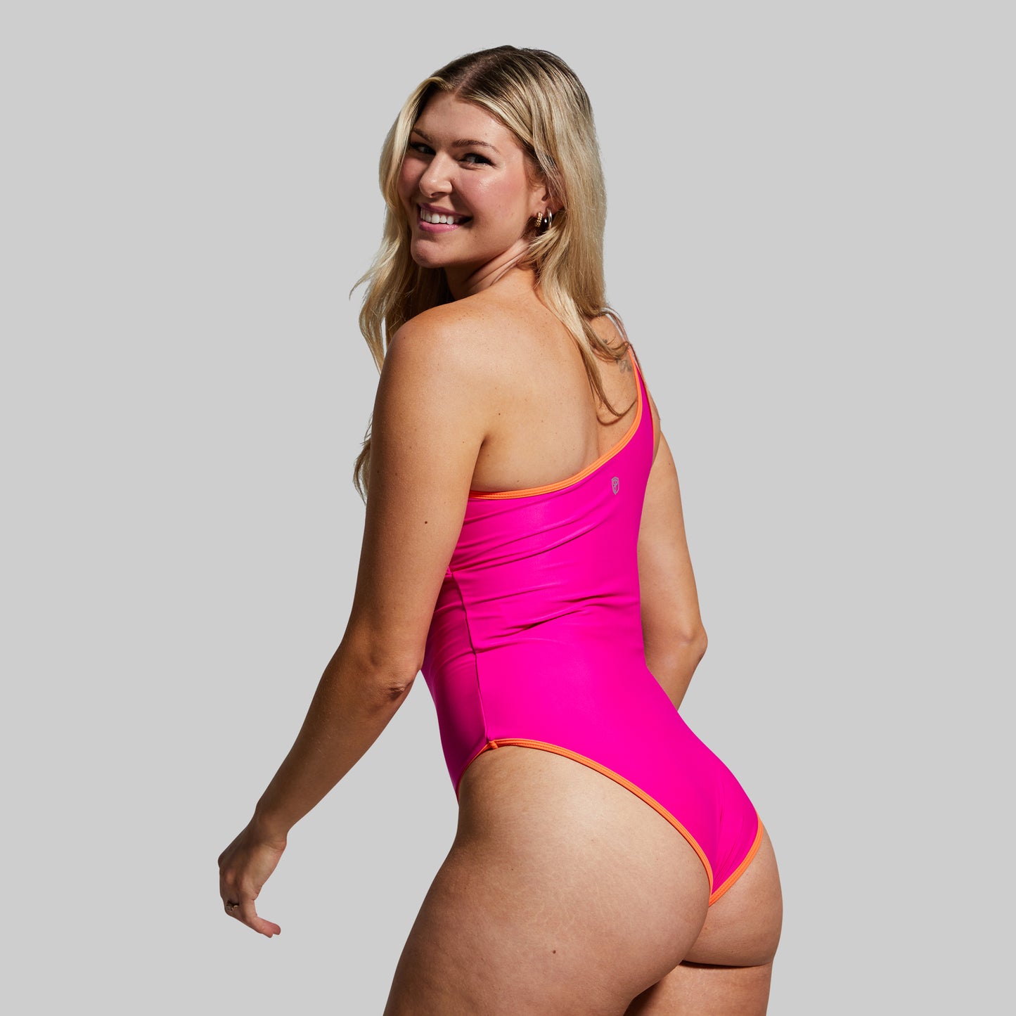 Back view of a woman wearing a bright pink one-shoulder swimsuit against a gray background