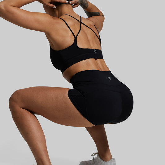 Rear view of a woman in a black sports bra and black high-waisted athletic shorts, crouching in a gym pose against a solid light gray background.