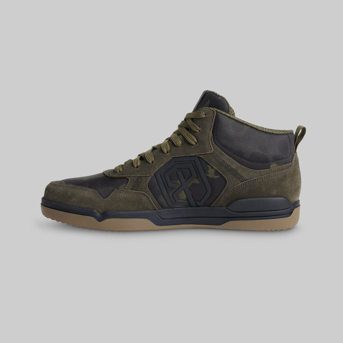A mid-top sneaker in olive green suede and dark camouflage panels, featuring a gum-colored sole, shown from a side profile on a light gray background.