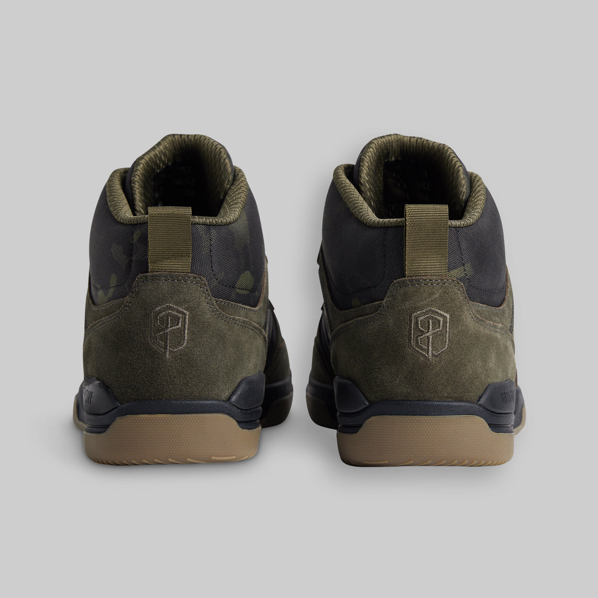 A pair of mid-top sneakers shown from the back, featuring olive green suede and camo panels, dark navy overlays, and tan rubber soles, set against a solid gray background.