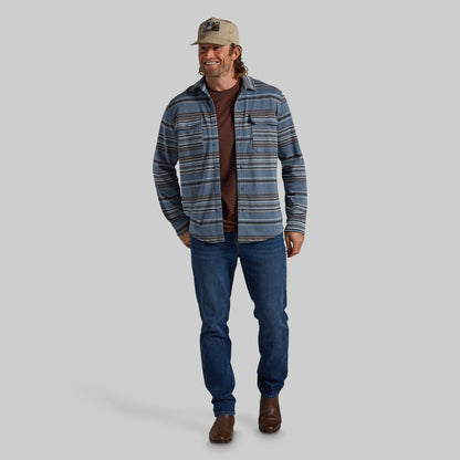 A man modeling a blue and gray striped flannel shirt over a brown t-shirt, paired with blue jeans, brown boots, and a beige baseball cap, against a solid gray background.