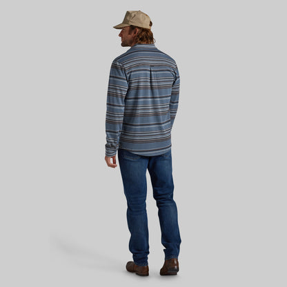A man shown from the back wearing a blue and gray striped long-sleeve shirt, dark blue jeans, brown shoes, and a beige cap, against a solid gray background.
