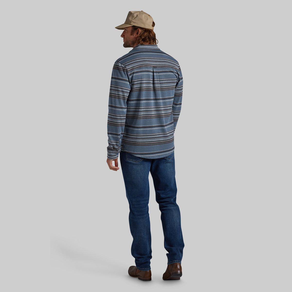 A man shown from the back wearing a blue and gray striped long-sleeve shirt, dark blue jeans, brown shoes, and a beige cap, against a solid gray background.
