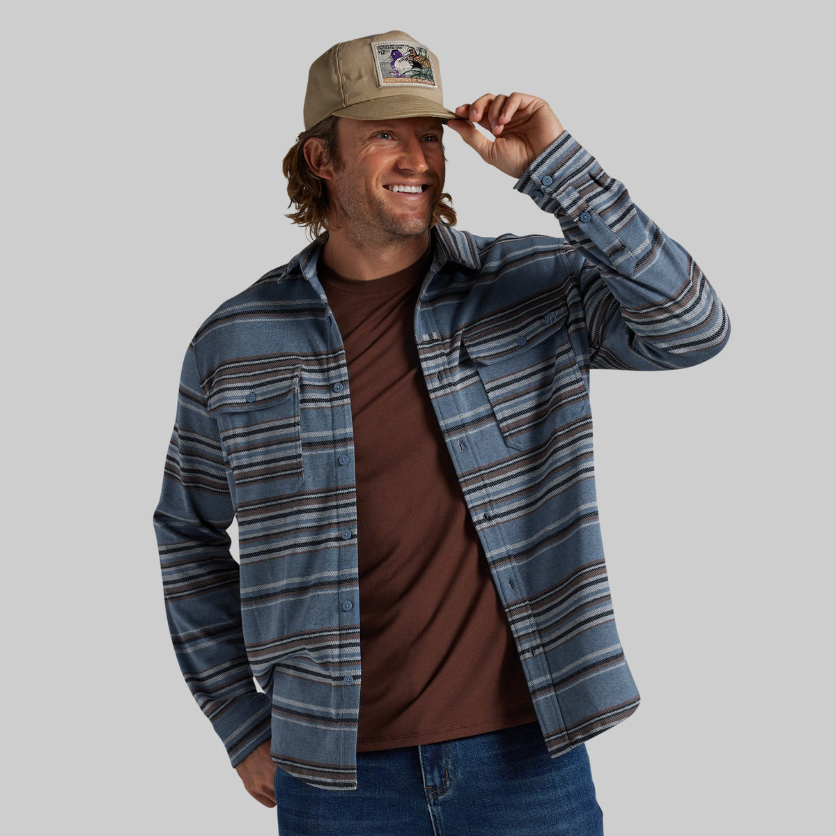 A man wearing a blue and gray striped flannel shirt jacket over a brown t-shirt, blue jeans, and a khaki baseball cap, posing against a solid gray background.