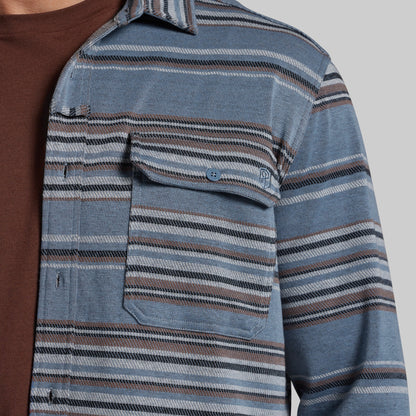 Close-up of a person wearing a blue striped button-up shirt over a brown t-shirt, showing the chest pocket and fabric texture against a light gray background.