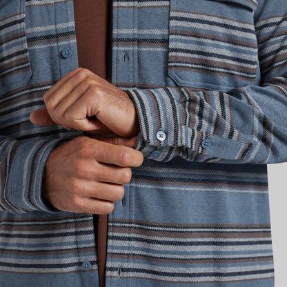 Close-up of a person wearing a blue striped button-up shirt, adjusting the cuff with both hands. The shirt features horizontal stripes in blue, gray, and brown, with a solid brown undershirt visible underneath.