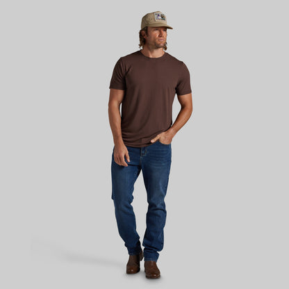 A man modeling a dark brown crewneck t-shirt, blue jeans, brown boots, and a beige baseball cap against a solid gray background.