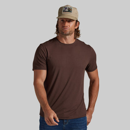 A man wearing a plain brown short-sleeve crewneck t-shirt and a beige trucker hat, standing against a solid gray background.