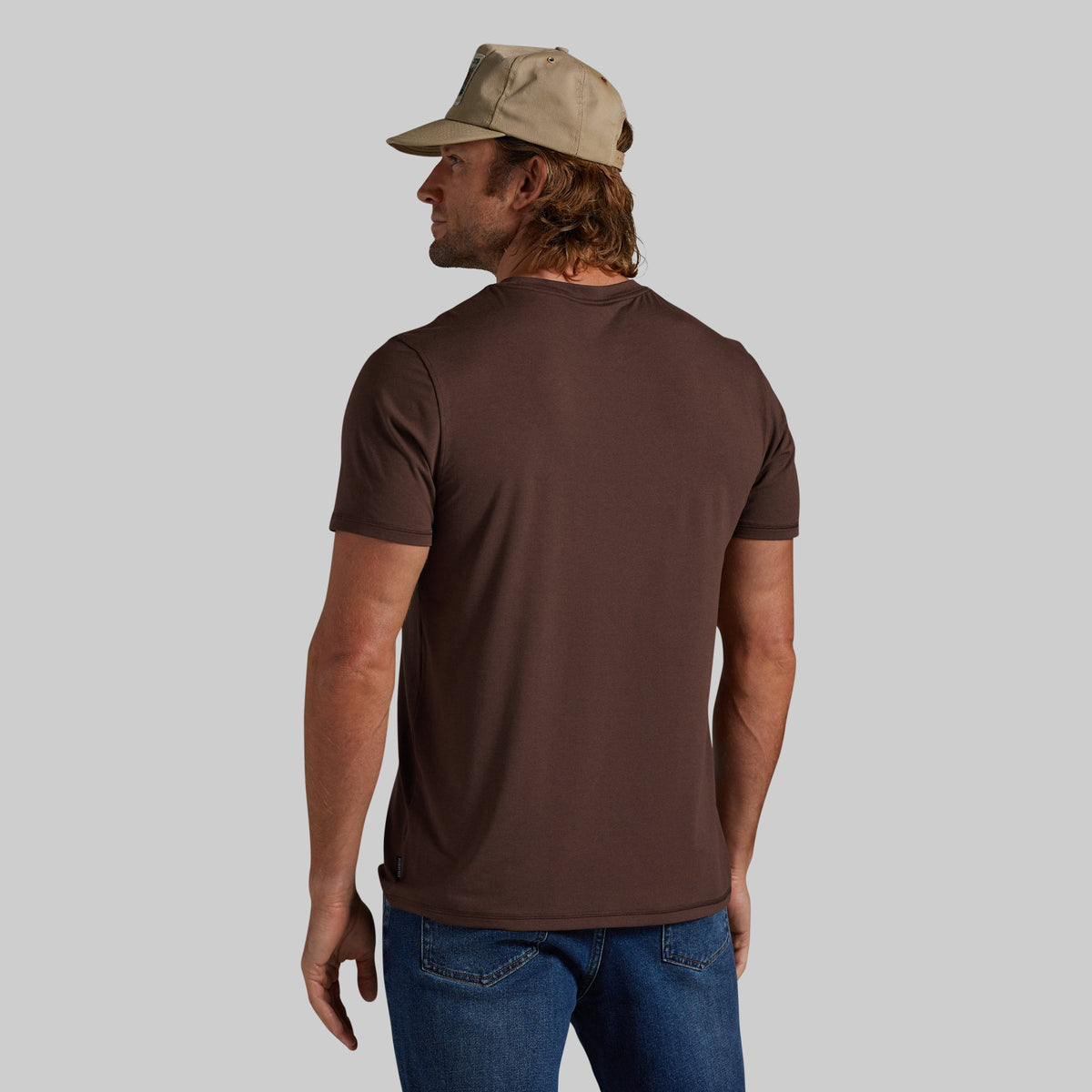 A man wearing a brown short-sleeve crewneck t-shirt, blue jeans, and a beige baseball cap, shown from the back against a solid gray background.