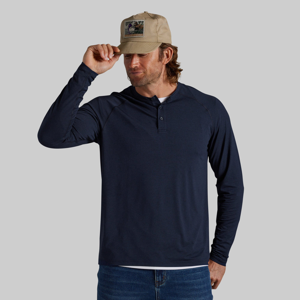 A man wearing a navy blue long-sleeve henley shirt, blue jeans, and a khaki baseball cap, standing against a solid gray background.
