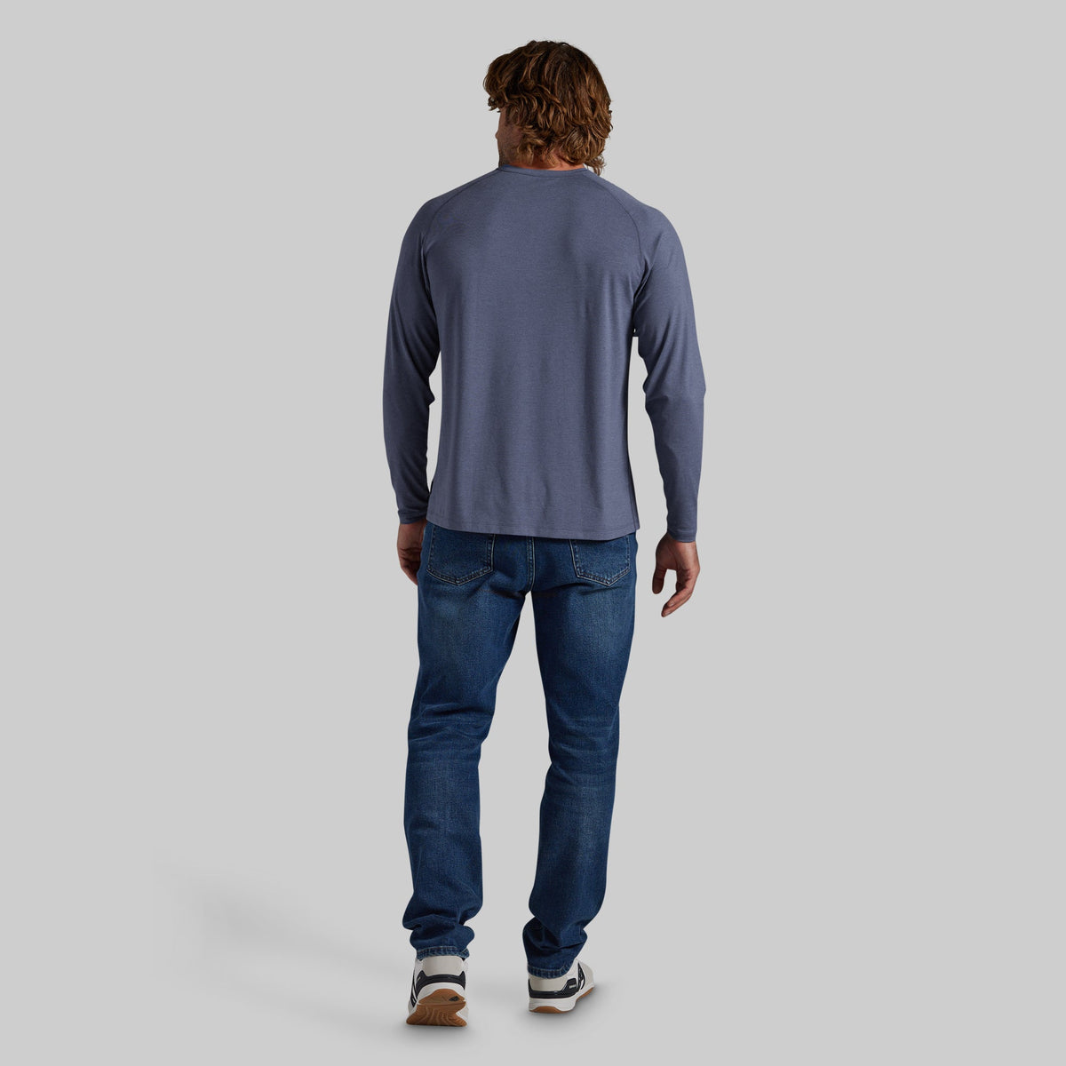 A man viewed from behind wearing a slate blue long-sleeve shirt and blue jeans, standing against a solid gray background.
