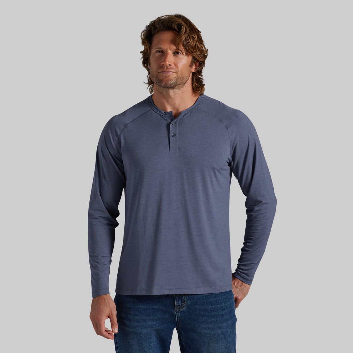 A man wearing a heathered slate blue long-sleeve henley shirt with blue jeans, standing against a solid gray studio background.