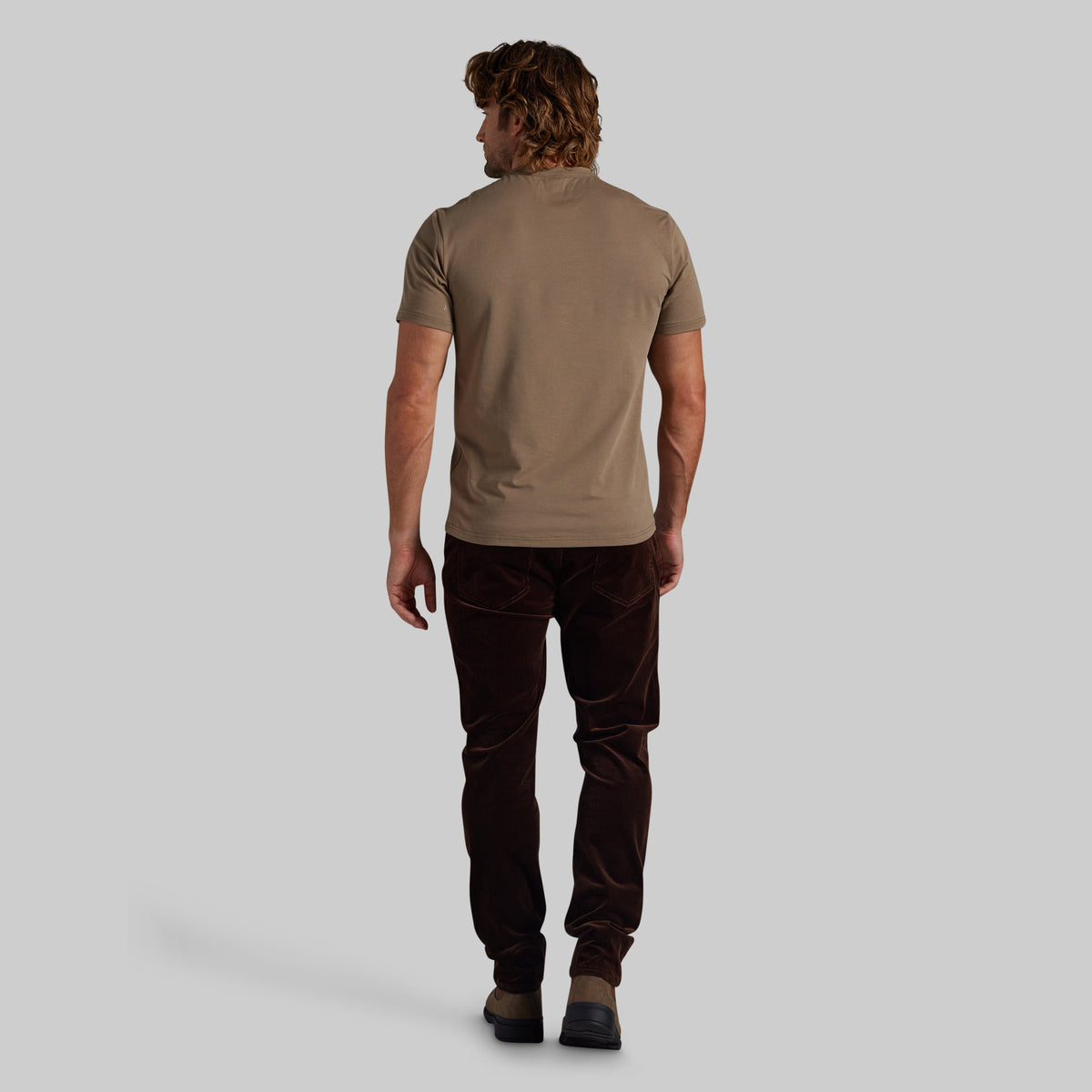 A man shown from behind wearing a short-sleeve taupe t-shirt and dark brown corduroy trousers, standing against a solid light gray background.