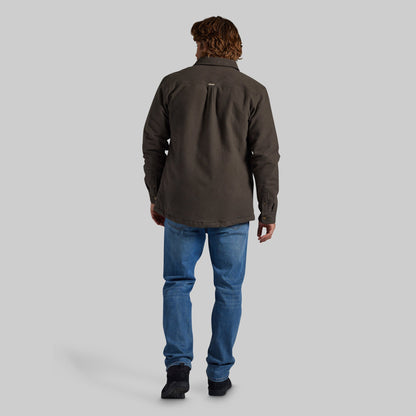 A man viewed from behind wearing a dark brown long-sleeve button-up shirt, blue jeans, and black shoes, standing against a solid gray studio background.