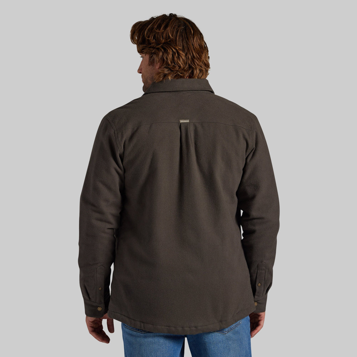 A man viewed from the back wearing a dark brown jacket with a vertical pleat on the back, paired with blue jeans, against a solid gray background.