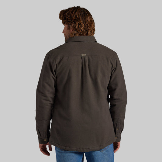 A man viewed from the back wearing a dark brown jacket with a vertical pleat on the back, paired with blue jeans, against a solid gray background.