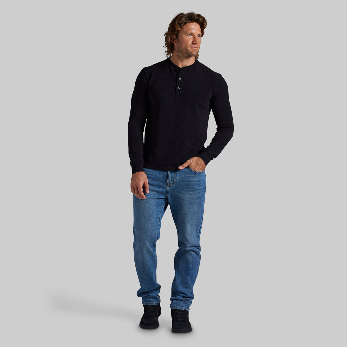 A man modeling a black long-sleeve henley shirt with blue jeans and black shoes, standing against a solid gray studio background.