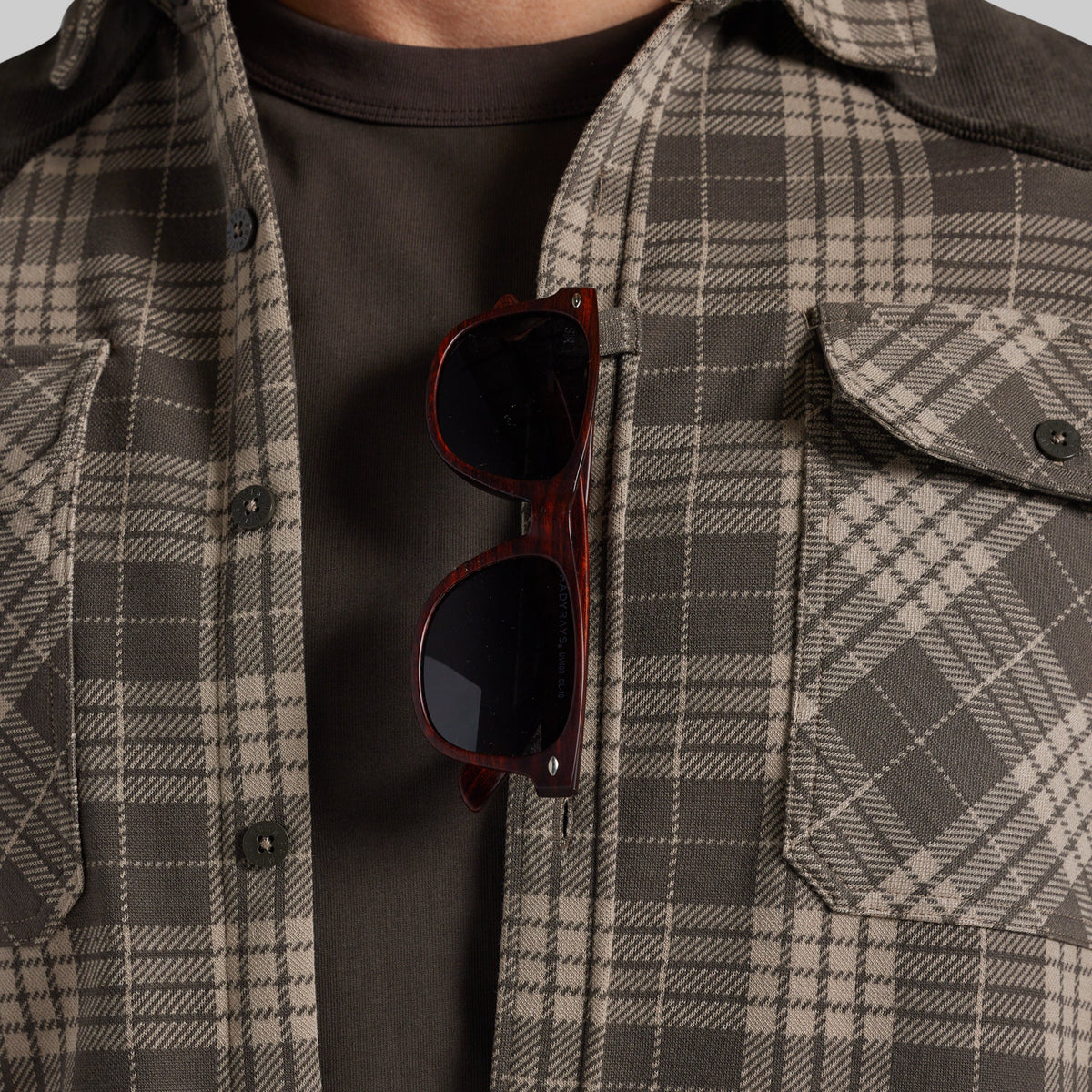 A close-up of a person wearing a brown and beige plaid overshirt over a dark brown t-shirt, with tortoiseshell sunglasses hanging from the placket.