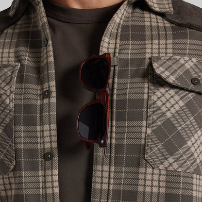 A close-up of a person wearing a brown and beige plaid overshirt over a dark brown t-shirt, with tortoiseshell sunglasses hanging from the placket.