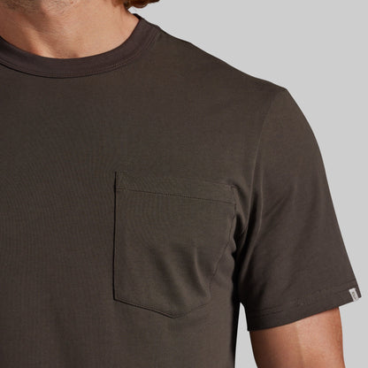 Close-up of a man wearing a dark brown short-sleeve crewneck t-shirt with a chest pocket, set against a solid gray background.