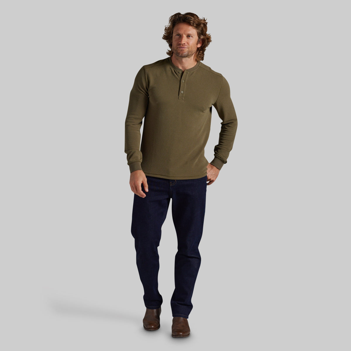 A man modeling a long-sleeve olive green henley shirt with dark navy pants and brown shoes, standing against a solid gray studio background.