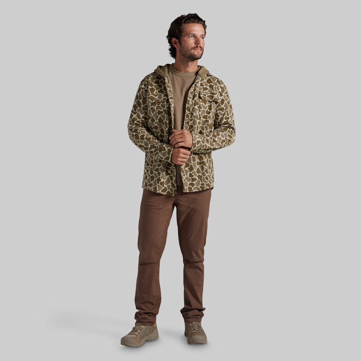 A man modeling a hooded flannel with a brown and beige animal print pattern, worn over a brown t-shirt and brown pants, against a solid gray background.