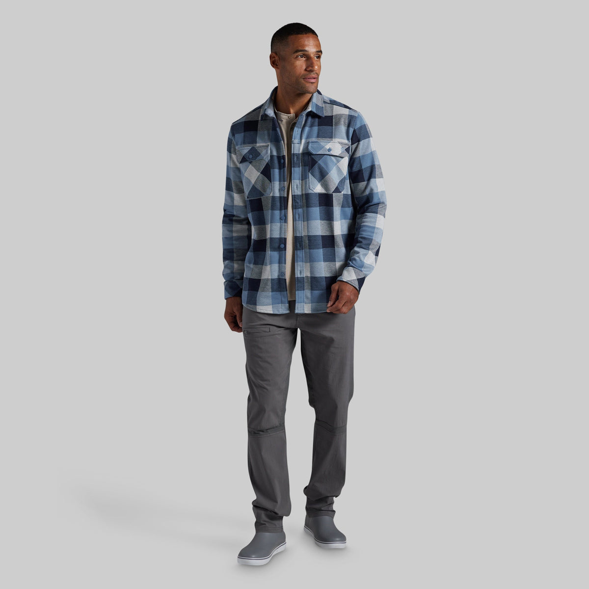 A man modeling a blue and gray plaid button-up flannel shirt, paired with dark gray pants and gray sneakers, against a solid light gray background.