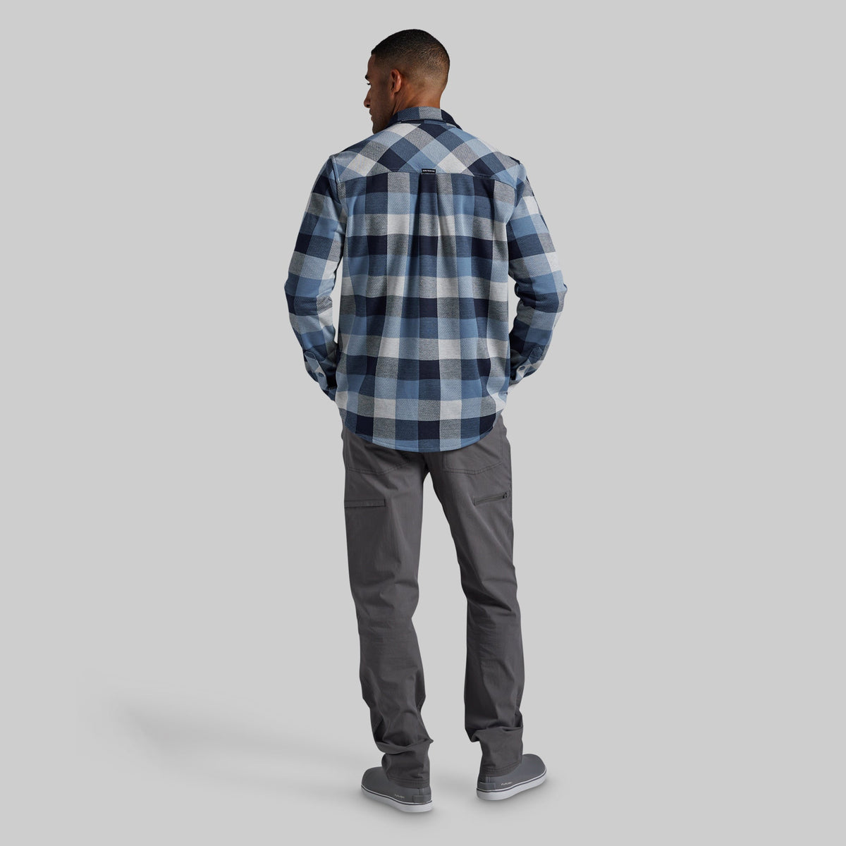 A man is shown from behind wearing a blue and white plaid flannel shirt and dark gray pants, standing against a solid gray studio background.