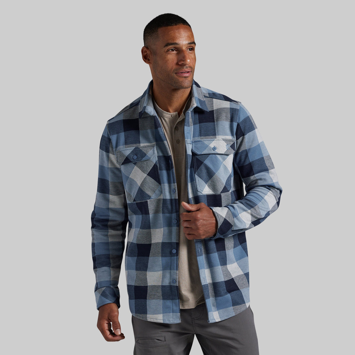 A man wearing a blue and navy plaid button-up flannel shirt over a beige t-shirt and gray pants, standing against a solid gray studio background.