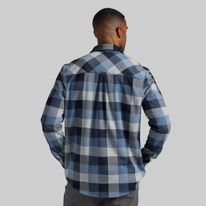 A man viewed from behind wearing a long-sleeve plaid flannel shirt in shades of navy, blue, gray, and white, paired with gray pants, against a solid gray background.