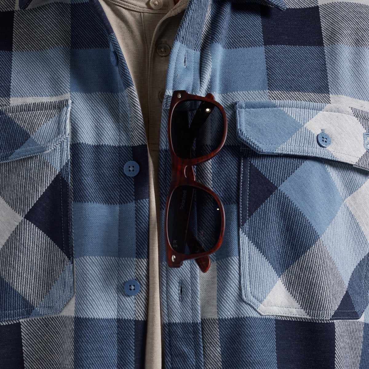 A close-up of a blue and navy plaid flannel button-up shirt with a pair of dark brown sunglasses hanging from the sunglass loop, layered over a beige undershirt.