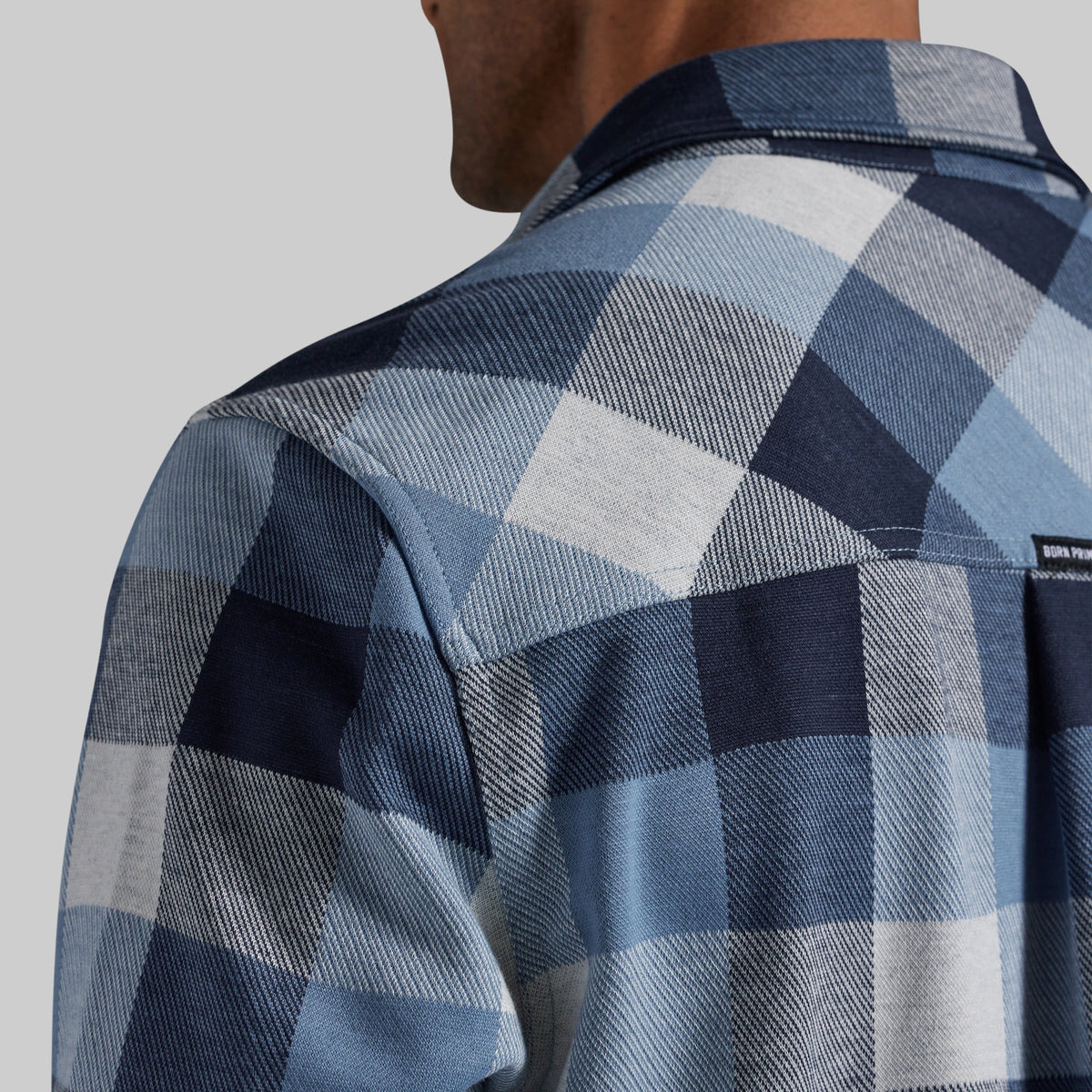 Close-up of a man wearing a blue and white plaid flannel shirt, showing the back of the neck, collar, and shoulder with a small visible brand tag.