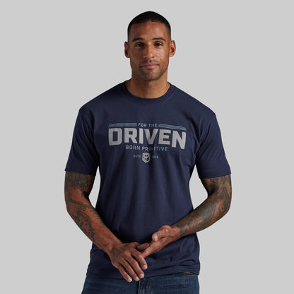 A man wearing a navy blue t-shirt with a distressed white graphic that reads 'FOR THE DRIVEN BORN PRIMITIVE' and a shield logo, set against a solid gray background.