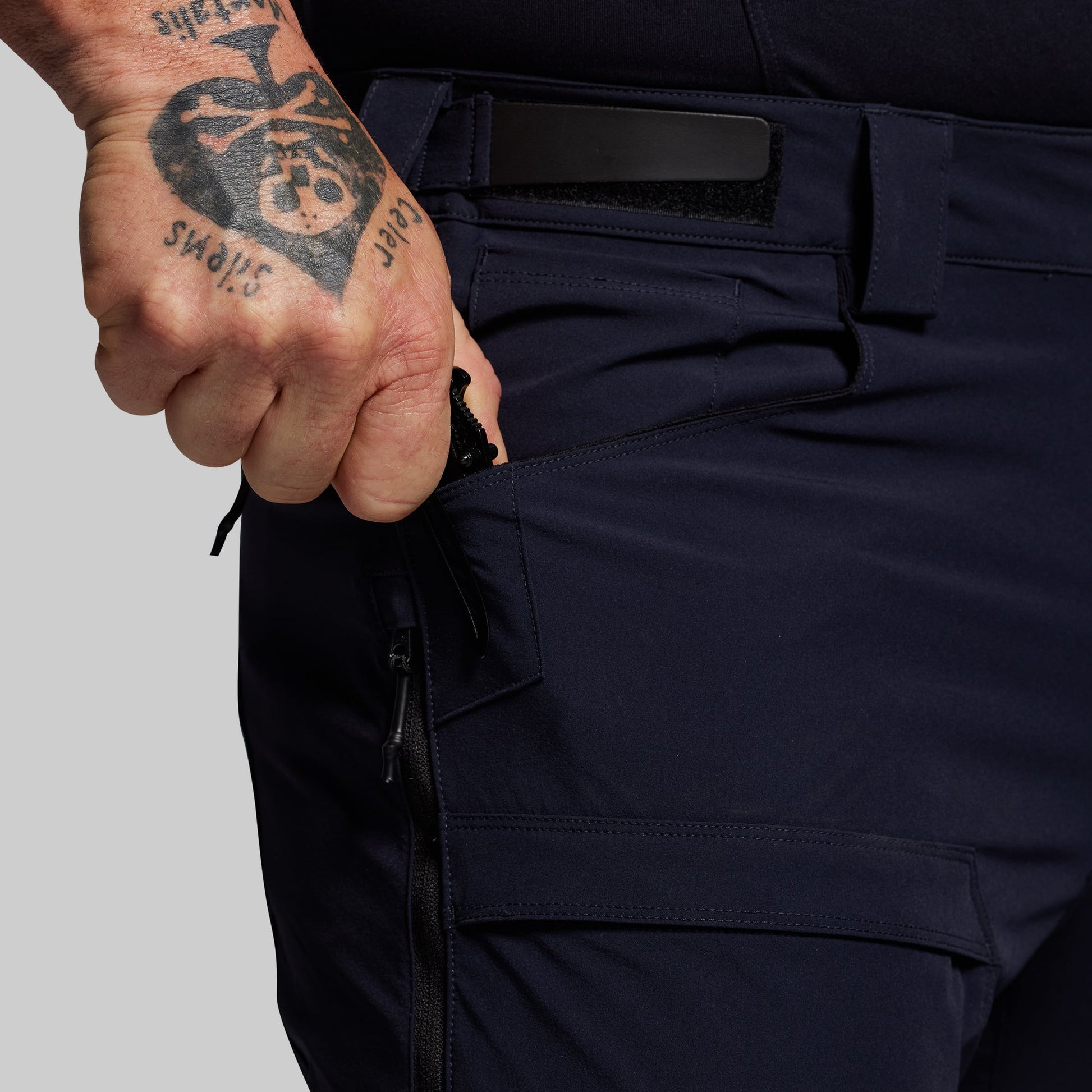 Person wearing dark blue outdoor pants with a tattooed hand on a gray background