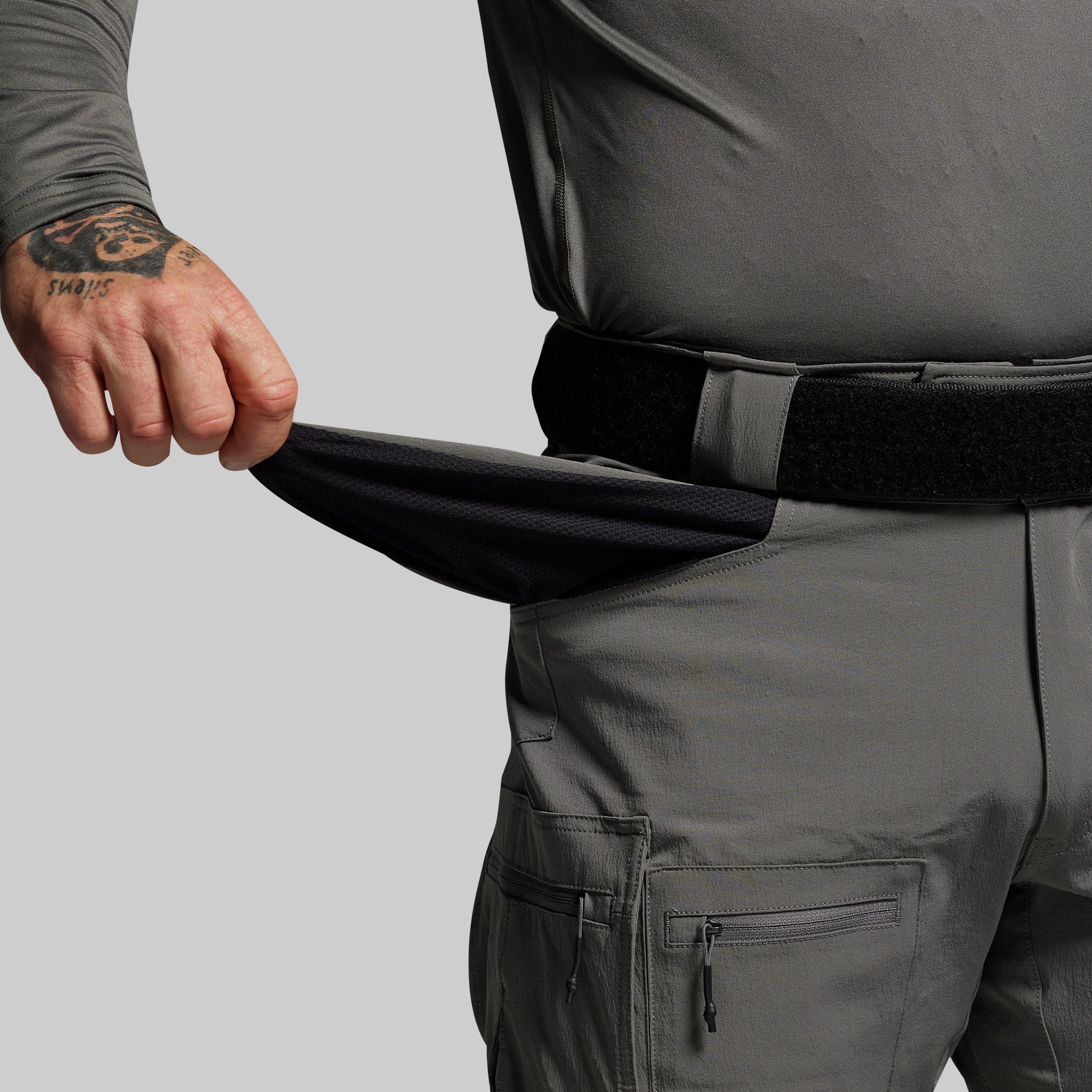 Person wearing dark gray pants with a tattooed arm on a light gray background