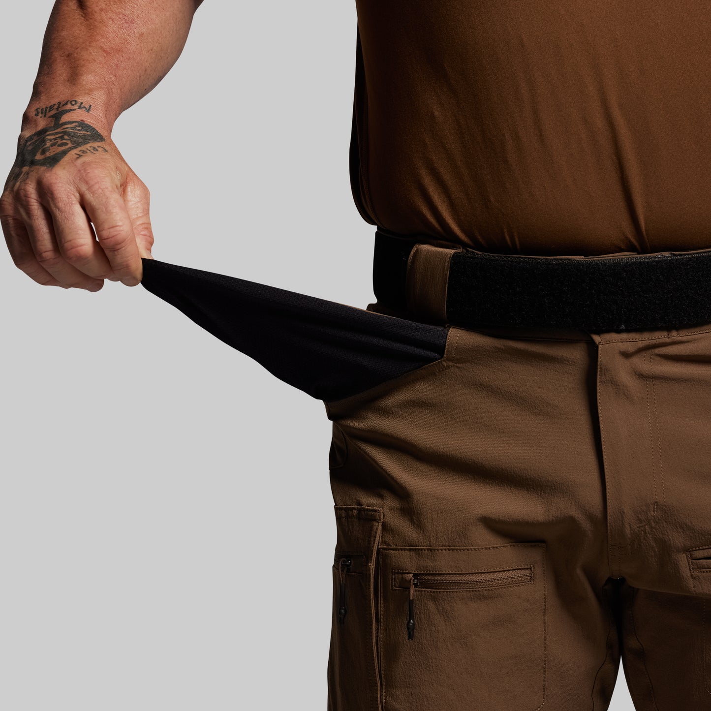 Person wearing brown pants with a black belt, showing the inside of front pocket on a gray background 