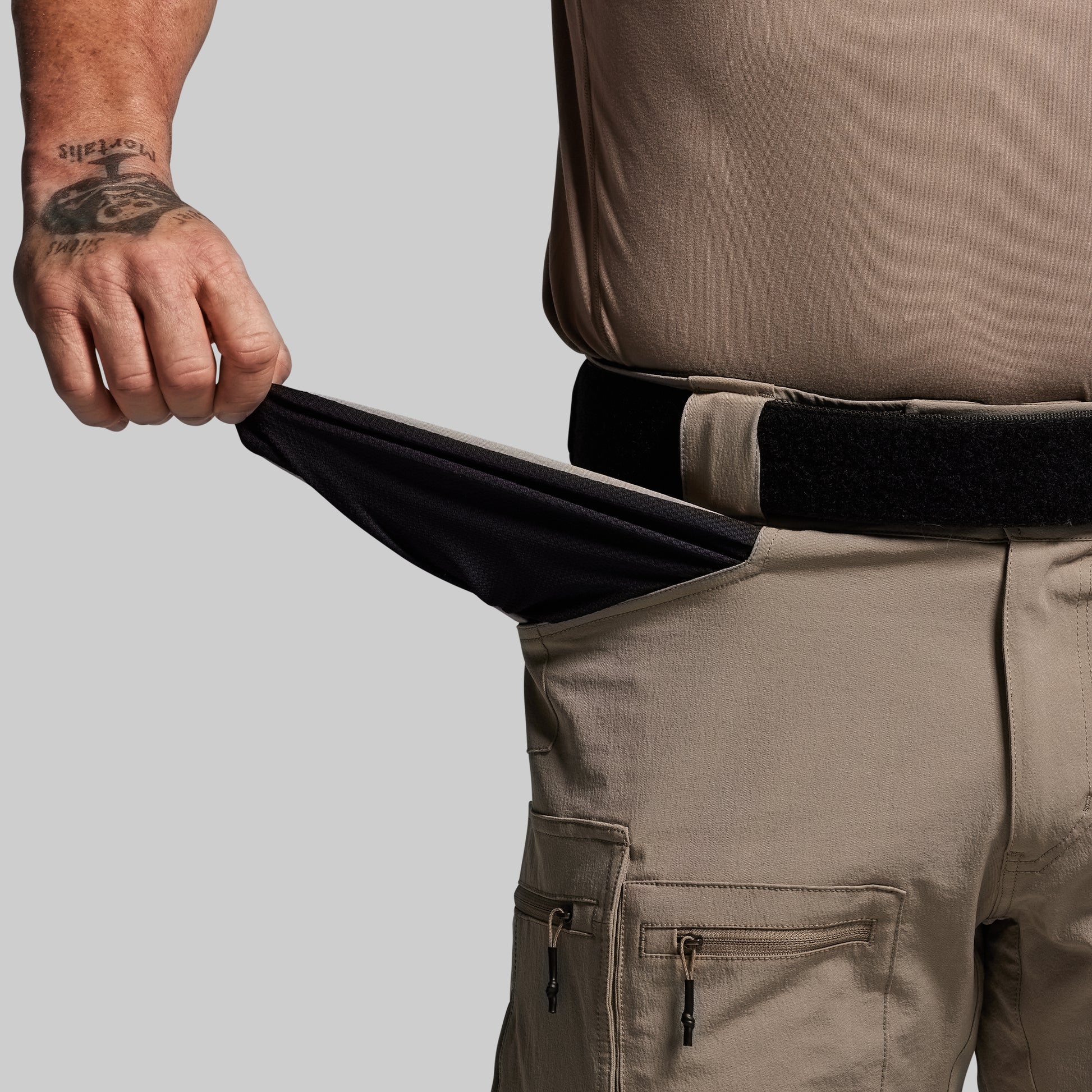 Person wearing tan pants with a black belt, showing the inside of front pocket on a gray background 