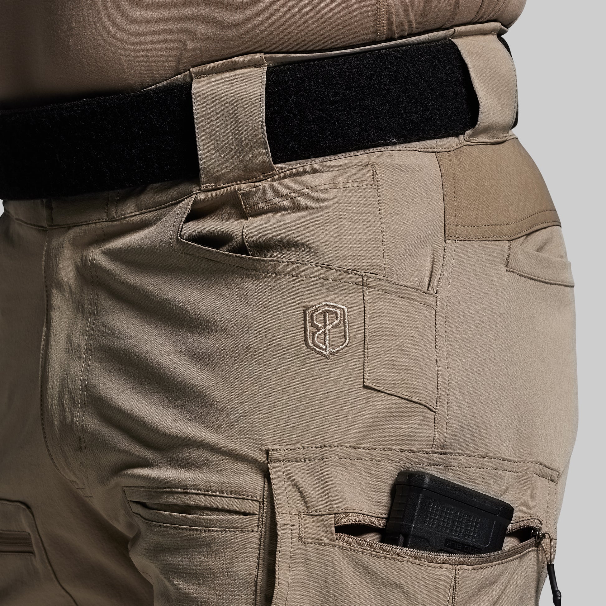 Tan tactical pants with a logo on the pocket against a gray background