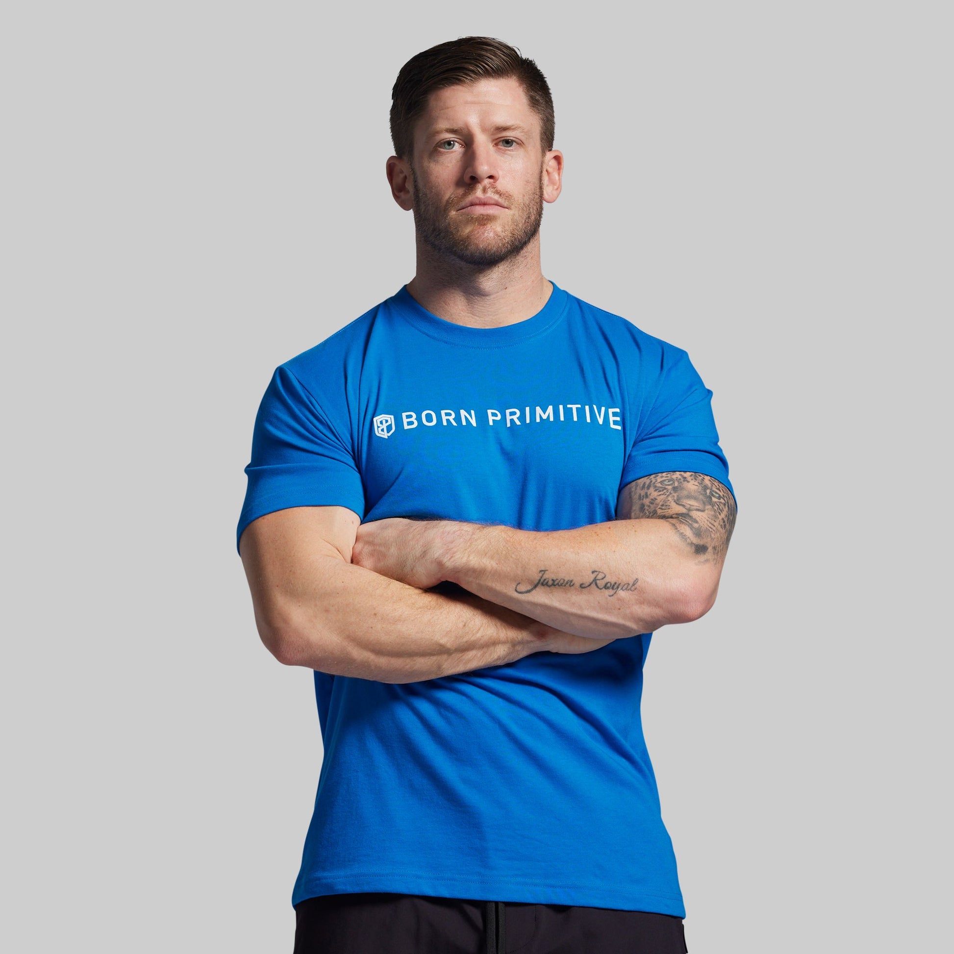 Man wearing a blue 'Born Primitive' t-shirt with arms crossed on a gray background