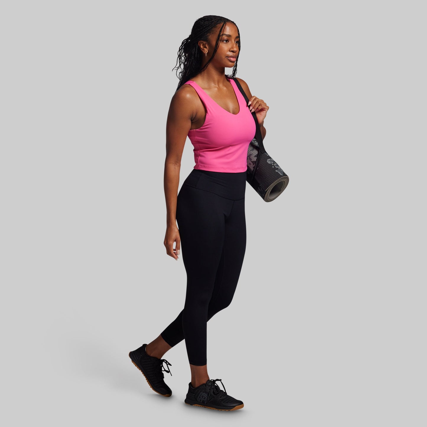Woman in pink top and black leggings holding a yoga mat on a gray background