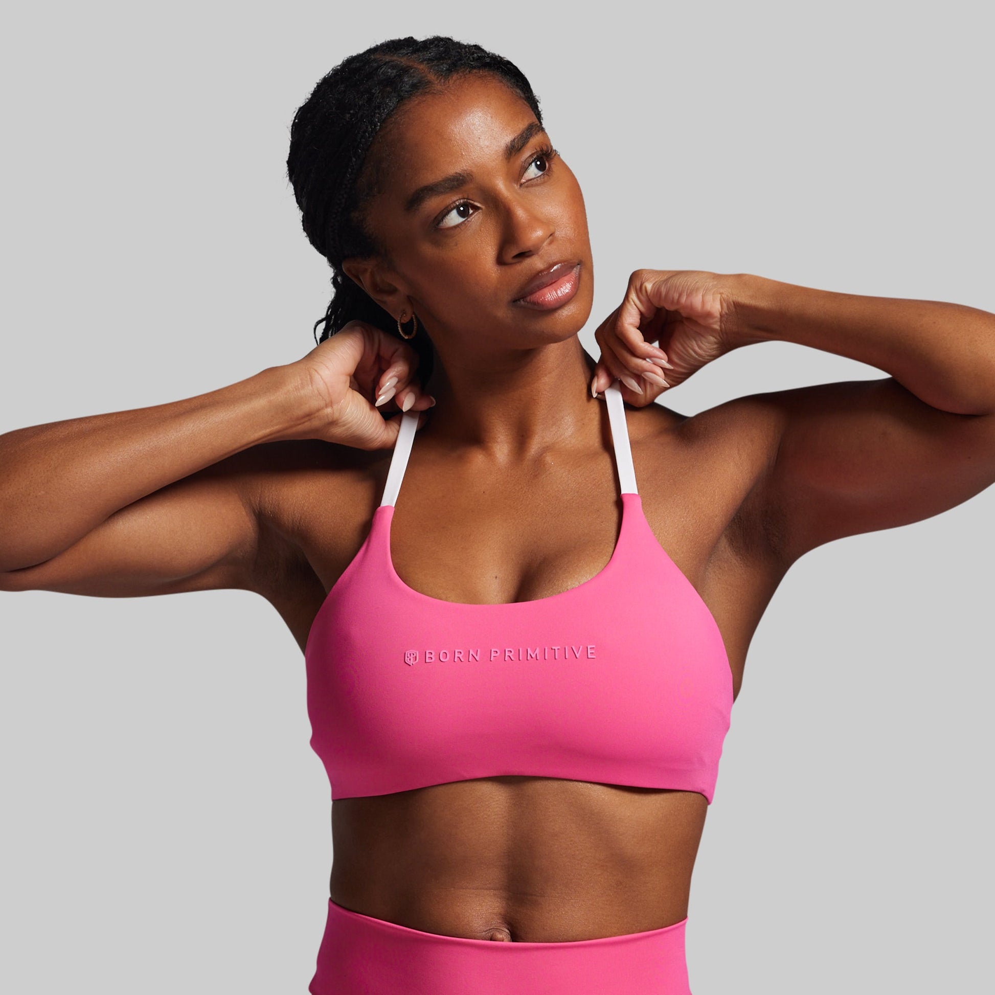 Woman wearing a pink sports bra with 'BORN PRIMITIVE' branding on a gray background